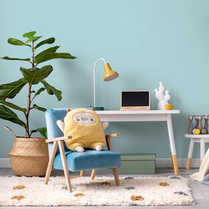 May include: A blue armchair with a yellow plush toy sitting on it. The chair is in a room with a white desk, a lamp, and a potted plant.