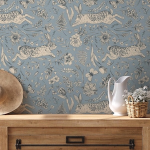 May include: A light blue wallpaper featuring a repeating pattern of running rabbits, flowers, and butterflies in beige and gray. A wooden table holds a straw hat, a white pitcher, and a wicker basket with white flowers.