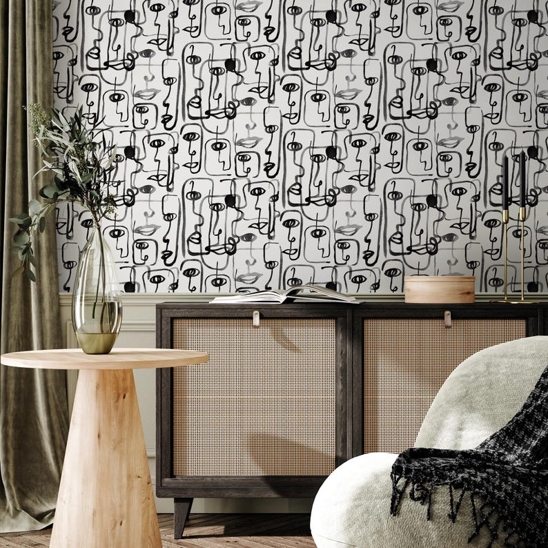 May include: Black and white abstract wallpaper with a repeating pattern of stylized faces. The wallpaper is shown on a wall in a room with a wooden table, a chair, and a cabinet.