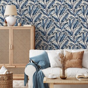 Op de afbeelding: Een kamer met een witte bank, blauwe en bruine kussens, een houten kast en een rieten mand. Het behang heeft een blauw bladerpatroon. Een lamp met een beige kap staat op de kast.