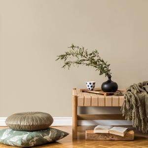 May include: A serene interior scene features a wooden bench with a woven seat, holding a black vase with greenery and a blue and white polka-dot cup. Two decorative pillows and a green throw blanket add texture and color. An open book rests on a wooden block.