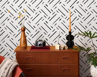 May include: A wooden dresser with three drawers holds a vintage camera, books, and decorative items. A tall, brown vase with yellow flowers and a black candle holder with a lit candle sit on top. The background features a white wall with a black geometric pattern.