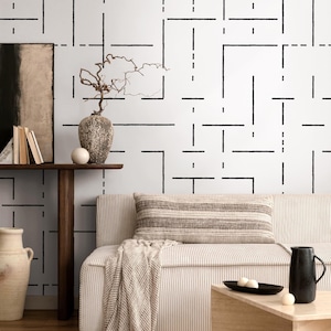 May include: A modern living room scene featuring a white sofa with a striped pillow and a beige throw. The wall has a black geometric pattern. A wooden side table holds a vase, books, and a decorative sphere. A black pitcher and tray are on a wooden coffee table.