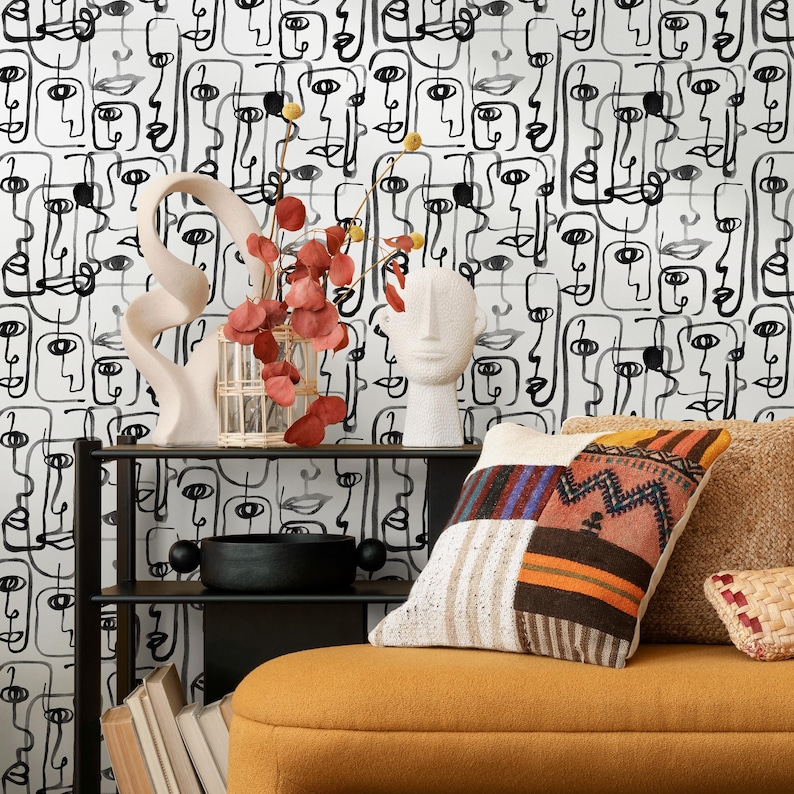 May include: A black and white abstract face pattern wallpaper with a white ceramic sculpture, a glass vase with red flowers, and a black metal shelf with a black bowl and books. The shelf is in front of a yellow couch with a patterned pillow.