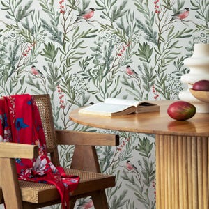 Op de afbeelding: Een witte achtergrond met een herhalend patroon van groene bladeren, rode bessen en roze vogels. Het patroon is op een muur achter een houten tafel met een boek en een vrucht erop.