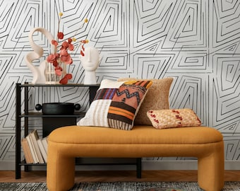 Può includere: Una panca giallo senape con tre cuscini decorativi davanti a una mensola nera. I cuscini hanno vari motivi e trame. Una scultura astratta bianca e un vaso con rami rossi sono sulla mensola. Lo sfondo è un muro bianco con un design geometrico nero.