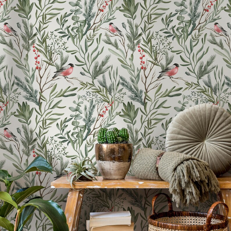 Op de afbeelding: Een witte behang met een herhalend patroon van groene bladeren, rode bessen en roze vogels. De behang wordt getoond in een kamer met een houten tafel, een cactus in een pot, een deken en een rond kussen.
