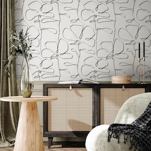 May include: A modern interior scene featuring a light-colored wall with a repeating black line art floral pattern. A wooden cabinet with woven panels sits below, and a round wooden side table is in the foreground. A vase with greenery and a gold candle holder are also visible.