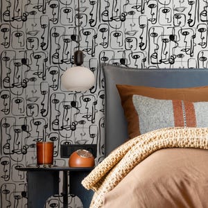 May include: A bedroom with a white and black patterned wallpaper featuring abstract faces. The bed has a brown duvet and a brown throw blanket. A bedside table with a lamp and a clock is next to the bed.