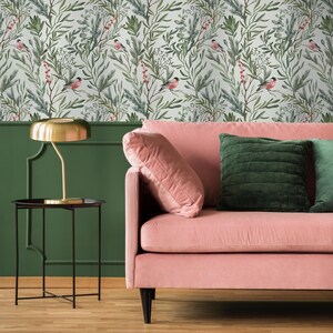 Op de afbeelding: Een roze fluwelen bank met groene fluwelen kussens staat voor een muur met een wit en groen bloemenbehang met kleine vogels. Een gouden tafellamp staat op een zwarte bijzettafel.