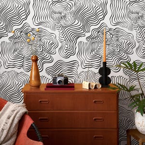 May include: A wooden dresser with three drawers holds a vintage camera, books, and decorative items. A tall, brown vase with yellow flowers and a black candle holder with an orange candle sit on top. The background features a black and white abstract pattern.