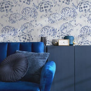 May include: A blue velvet sofa with two cushions, one round and one rectangular, in front of a wall with a blue and white floral wallpaper pattern. A dark blue cabinet with a gold clock and a blue vase sits on top of the cabinet.