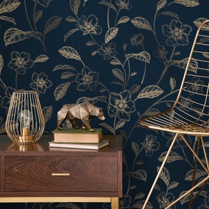 May include: A dark blue floral wallpaper with gold-colored flowers and leaves. A gold wire lamp with a lightbulb, a geometric bear sculpture, and a gold chair are in the foreground. A dark wood nightstand with a drawer is also visible.