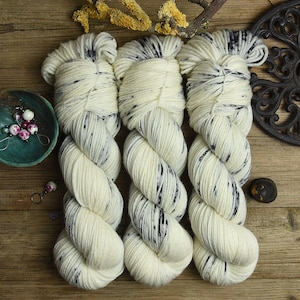 Op de afbeelding: Drie strengen crèmekleurig garen met donkerblauwe spikkels. Het garen is getwist en op een houten oppervlak geplaatst. Een kleine turquoise kom met sieraden en een decoratief metalen stuk zijn ook zichtbaar.