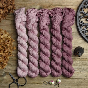 Könnte beinhalten: Fünf Garnknäuel in verschiedenen Rosa- und Mauve-Tönen sind auf einer Holzoberfläche angeordnet. Das Garn ist zu ordentlichen Bündeln gewickelt. Eine Schere, Trockenblumen und dekorative Gegenstände sind ebenfalls vorhanden.