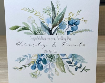 May include: A white wedding card with a watercolor illustration of blue and green flowers and leaves. The text reads "Congratulations on your Wedding Day Kirsty & Paul 24th May 2022".
