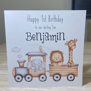 May include: A white card with a watercolor illustration of a train with an elephant, lion, and giraffe. The card says "Happy 1st Birthday to our darling Son Benjamin".