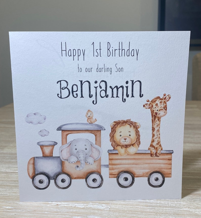 May include: A white card with a watercolor illustration of a train with an elephant, lion, and giraffe. The card says "Happy 1st Birthday to our darling Son Benjamin".