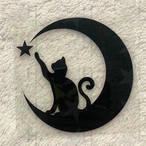 Könnte beinhalten: Schwarze Silhouette einer Katze, die nach einem Stern auf einem Halbmond greift. Das Design ist auf einem transparenten Hintergrund.