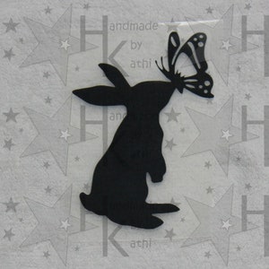 Könnte beinhalten: Schwarze Silhouette eines Kaninchens mit einem Schmetterling im Maul. Das Kaninchen steht auf seinen Hinterbeinen und hat seine Vorderpfoten erhoben. Der Schmetterling ist schwarz-weiß und hat eine Flügelspannweite von etwa 5 cm.