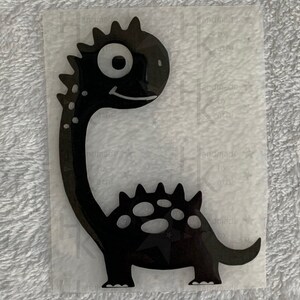 Könnte beinhalten: Schwarze Silhouette eines Cartoon-Dinosauriers mit einem großen Auge und einem Lächeln. Der Dinosaurier hat Stacheln auf dem Rücken und einen langen Hals.