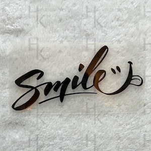 Könnte beinhalten: Schwarzer Vinyl-Aufkleber mit dem Wort "Smile" in einer stilisierten Schriftart mit einem Smiley am Ende.