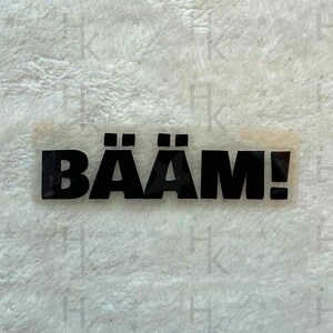 Könnte beinhalten: Ein weißer Aufkleber mit dem Text "BÄÄM!" in schwarzen Fettdruck.