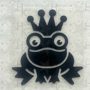 Könnte beinhalten: Schwarze Silhouette eines Frosches, der eine Krone trägt. Der Frosch sitzt mit gekreuzten Armen und hat große, runde Augen.