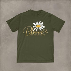 Puede incluir: Camiseta verde oliva con un diseño de margarita y la frase "Bloom where you are planted" en letras doradas. La margarita tiene pétalos blancos y un centro amarillo. Un pequeño corazón está al lado de la palabra "Bloom".