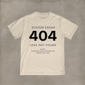 Könnte beinhalten: Ein beiges T-Shirt mit einer schwarzen Grafik, auf der "SYSTEM ERROR 404 LOVE NOT FOUND" und "SORRY! CURRENTLY BUSY LOOKING FOR MONEY, NOT LOVE" steht.