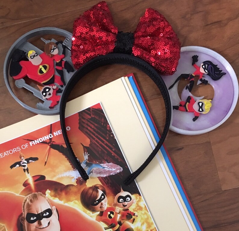 May include: A pair of black mouse ears with a red sequined bow. The ears are decorated with 3D figures of the Incredibles family from the Disney Pixar movie.