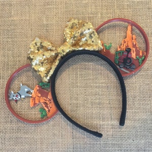 May include: A pair of brown and black mouse ears with a gold sequined bow. The ears feature a 3D design of a train and a mountain range on one side and a cartoon dog on the other.