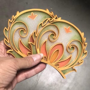 May include: Two decorative, layered coasters with an ornate floral design. The coasters feature a combination of colors, including orange, green, and yellow. The design includes swirling patterns and a stylized flower shape.