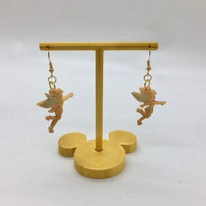 Puede incluir: Un par de pendientes de aro dorados con una figura de hada rosa claro con detalles de purpurina. Los pendientes se muestran en un soporte dorado.