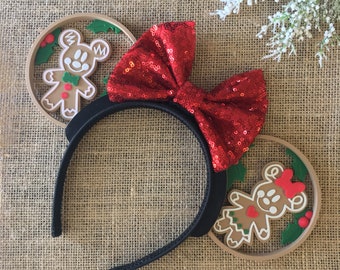 May include: A pair of black mouse ears with a red sequined bow. Each ear features a gingerbread Mickey Mouse character inside a brown circle with green holly leaves.
