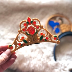 Puede incluir: Una tiara de plástico dorada con una rosa roja y hojas verdes. La tiara tiene joyas rojas y verdes y un clip negro en la parte posterior.
