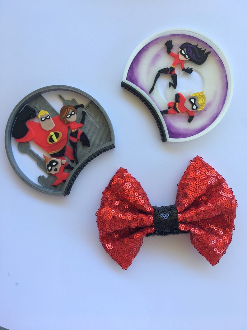 May include: A pair of gray and purple mouse ears with a red bow. The mouse ears feature a 3D image of the Incredibles family. The bow is made of red sequins with a black center.