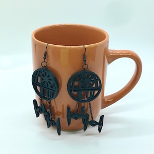 May include: A pair of black dangle earrings featuring a Death Star design with a smaller X-Wing fighter design hanging below. The earrings are displayed on a brown mug.