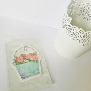 May include: A floral-themed sticker featuring a watercolor-style basket filled with pink roses and berries. The basket is light blue with a watercolor effect. The sticker is in a clear plastic sleeve. The text "Beautiful Flowers" is at the bottom.