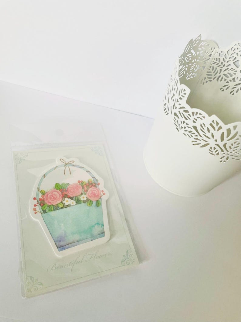 May include: A floral-themed sticker featuring a watercolor-style basket filled with pink roses and berries. The basket is light blue with a watercolor effect. The sticker is in a clear plastic sleeve. The text "Beautiful Flowers" is at the bottom.