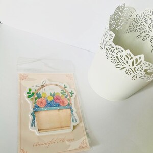 May include: A sticker featuring a watercolor illustration of a basket filled with colorful flowers, including pink roses and yellow blooms. The sticker is on a pink card with the text "Beautiful Flowers". A white, decorative metal container is also in the image.
