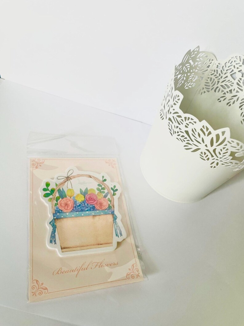 May include: A sticker featuring a watercolor illustration of a basket filled with colorful flowers, including pink roses and yellow blooms. The sticker is on a pink card with the text "Beautiful Flowers". A white, decorative metal container is also in the image.