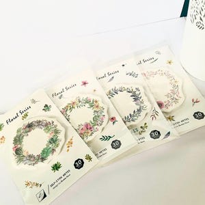 Könnte beinhalten: Vier Packungen mit selbstklebenden Notizen im floralen Design. Jede Packung enthält 30 Notizen und zeigt ein anderes Design mit einem floralen Kranz. Die Kränze sind in den Farben Grün, Lila, Rosa und Blau gehalten.