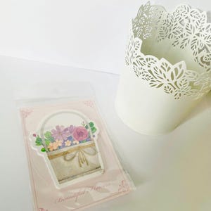 May include: A sticker featuring a watercolor illustration of a basket filled with purple, pink, and orange flowers. The sticker is on a pink card with the text "Beautiful Flowers". A white decorative pot is in the background.