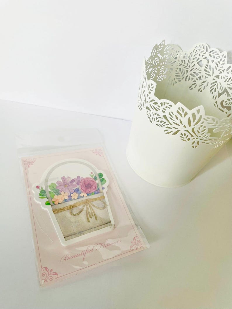 May include: A sticker featuring a watercolor illustration of a basket filled with purple, pink, and orange flowers. The sticker is on a pink card with the text "Beautiful Flowers". A white decorative pot is in the background.