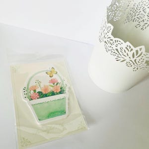 May include: A sticker featuring a watercolor illustration of a basket filled with pink and orange flowers and a yellow butterfly. The sticker is in a clear plastic sleeve, next to a white, decorative, cut-out metal pot.
