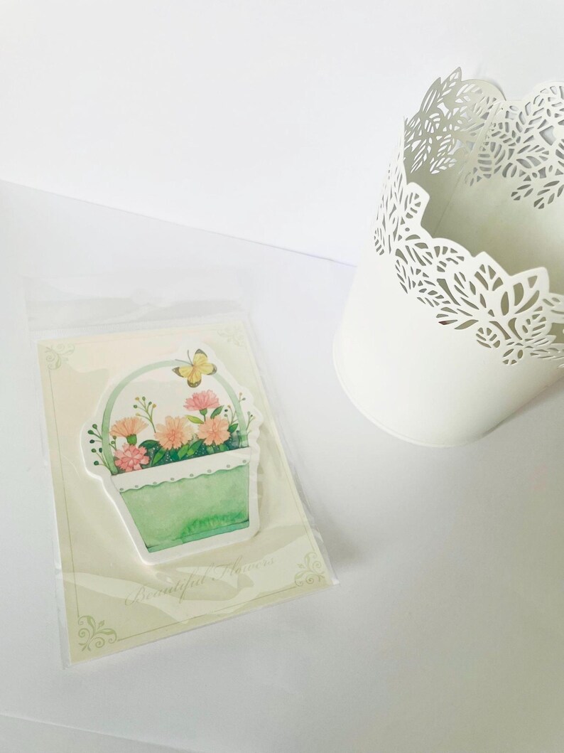 May include: A sticker featuring a watercolor illustration of a basket filled with pink and orange flowers and a yellow butterfly. The sticker is in a clear plastic sleeve, next to a white, decorative, cut-out metal pot.