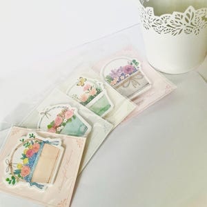 May include: A collection of floral-themed stickers in clear packaging. The stickers feature watercolor illustrations of flower arrangements in baskets and wreaths, with soft pastel colors and the text "Beautiful Flowers". A white decorative pot is in the background.