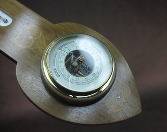 May include: A wooden barometer with a brass face and a glass cover. The face has a white background with black markings and a blue needle. The barometer reads "STORM" and "VERY DRY".