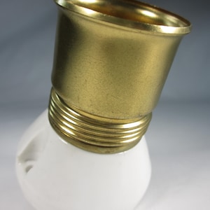 Puede incluir: Una base de bombilla de cerámica blanca con una parte superior de metal dorado. La parte superior tiene un anillo roscado y un borde liso y redondeado.